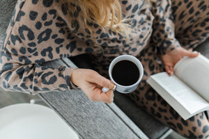 Woman relaxing on couch, drinking a coffee and reading while wearing a leopard print lounge set with long sleeves and pants.